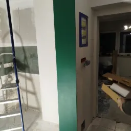 Wnętrze w trakcie prac: ściana z fragmentem pomalowanym na intensywny zielony kolor, widoczna drabina malarska, niezabezpieczone przewody elektryczne i rozdzielnia, ślady gładzi na podłodze.