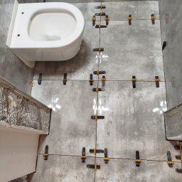 Krzysztof - Układanie szarych płytek podłogowych w łazience z zamontowaną białą miską WC, widoczne czarne krzyżyki dystansowe i żółte kliny poziomujące.