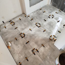 Krzysztof - Szare płytki podłogowe w trakcie układania, z użyciem klipsów poziomujących, widoczna puszka z instalacją wodną w ścianie i wiadro z zaprawą.