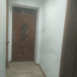 Krzysztof - Korytarz po remoncie z brązowymi panelami podłogowymi imitującymi drewno oraz brązowymi drzwiami wejściowymi z szybą z ozdobnym wzorem, ściany pomalowane na biało.