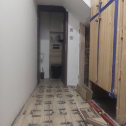 Krzysztof - Korytarz w trakcie kładzenia podłogi z płytek imitujących drewno, widoczne kliny dystansowe, płytki przygotowane do ułożenia, drzwi do łazienki w tle.