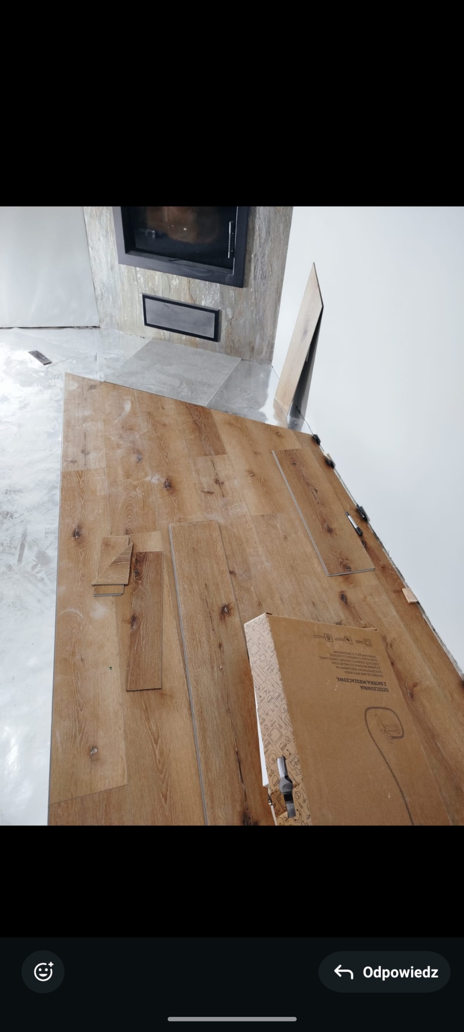 Fragment podłogi z widocznym kominkiem w trakcie układania paneli podłogowych o jasnym odcieniu drewna, z widocznym kartonem z nowymi panelami i przymiarem.