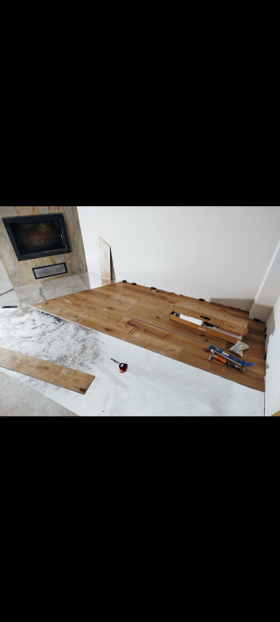 Fragment pokoju z kominkiem w trakcie układania podłogi z paneli drewnopodobnych, widoczne narzędzia i akcesoria montażowe.