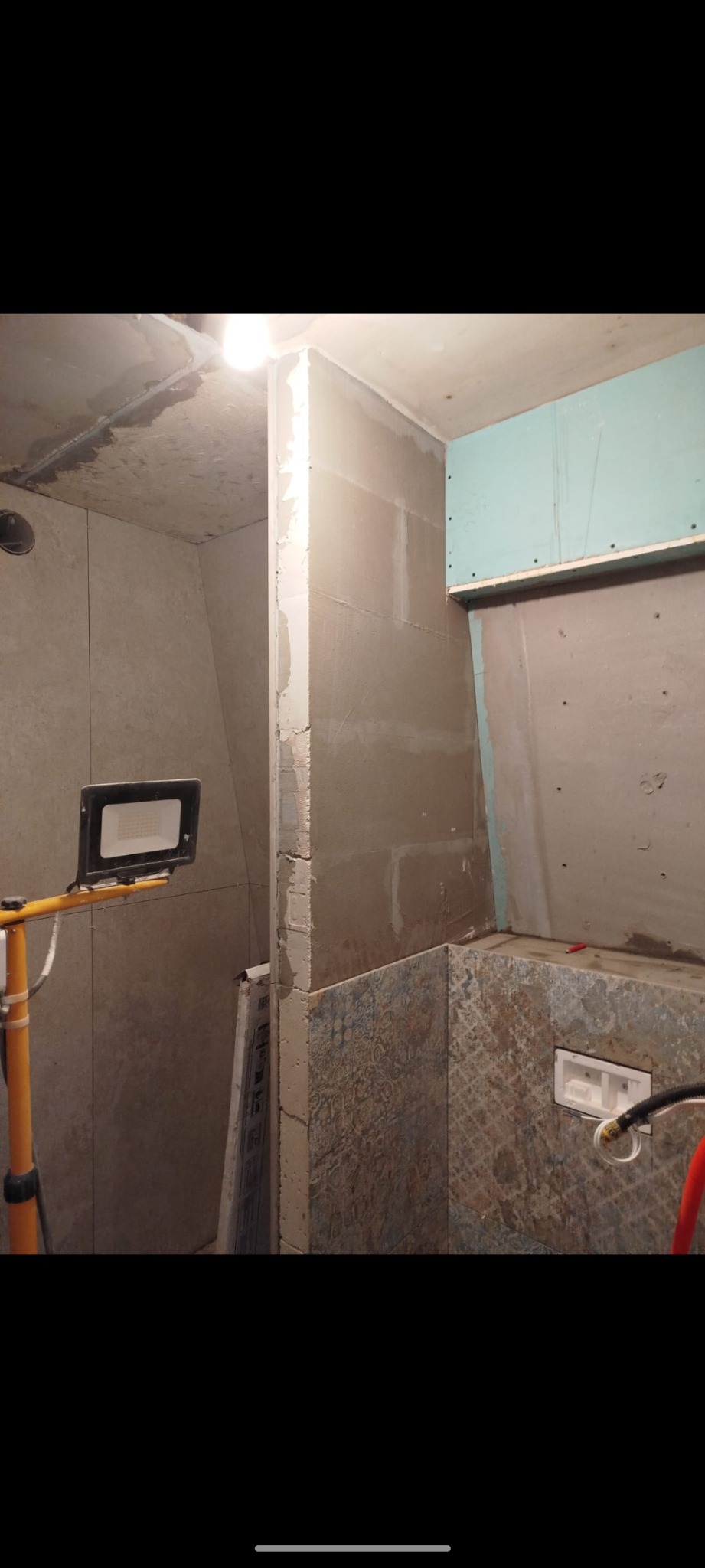 Wnętrze łazienki w trakcie remontu, widoczne płytki z wzorem, częściowo położone, oraz przygotowane ściany pod dalsze prace, oświetlone lampą roboczą.