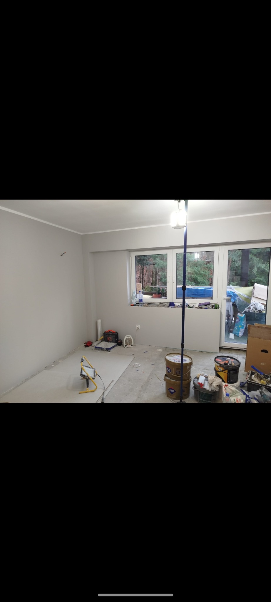 Pokój w trakcie prac remontowych, szare ściany, widoczne puszki z farbą, narzędzia malarskie, grzejnik elektryczny i lampa budowlana na wysięgniku.