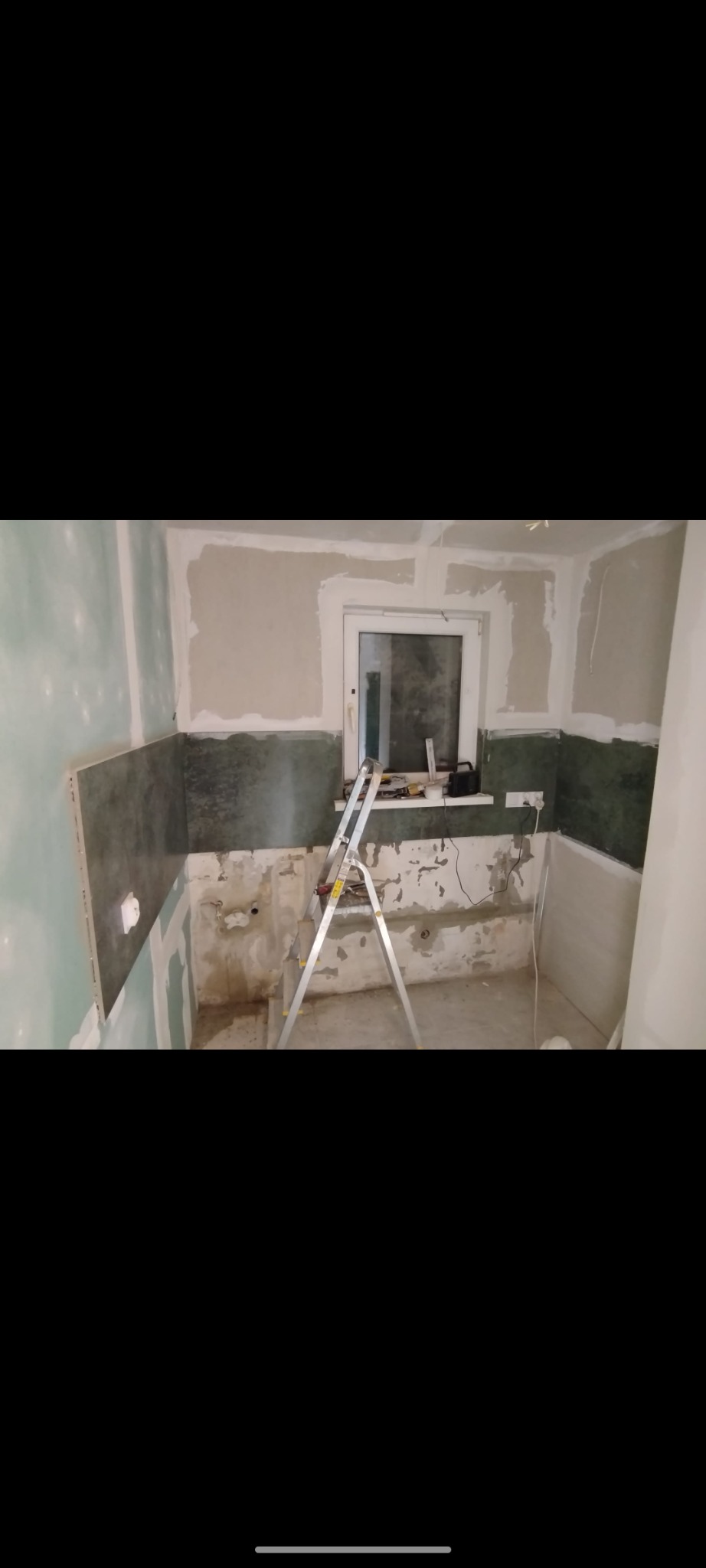 Fragment remontowanej kuchni z częściowo położonymi płytkami ściennymi w odcieniach zieleni i szarości, widoczne ślady po instalacjach, drabina aluminiowa, okno z parapetem, ściany przygotowane...