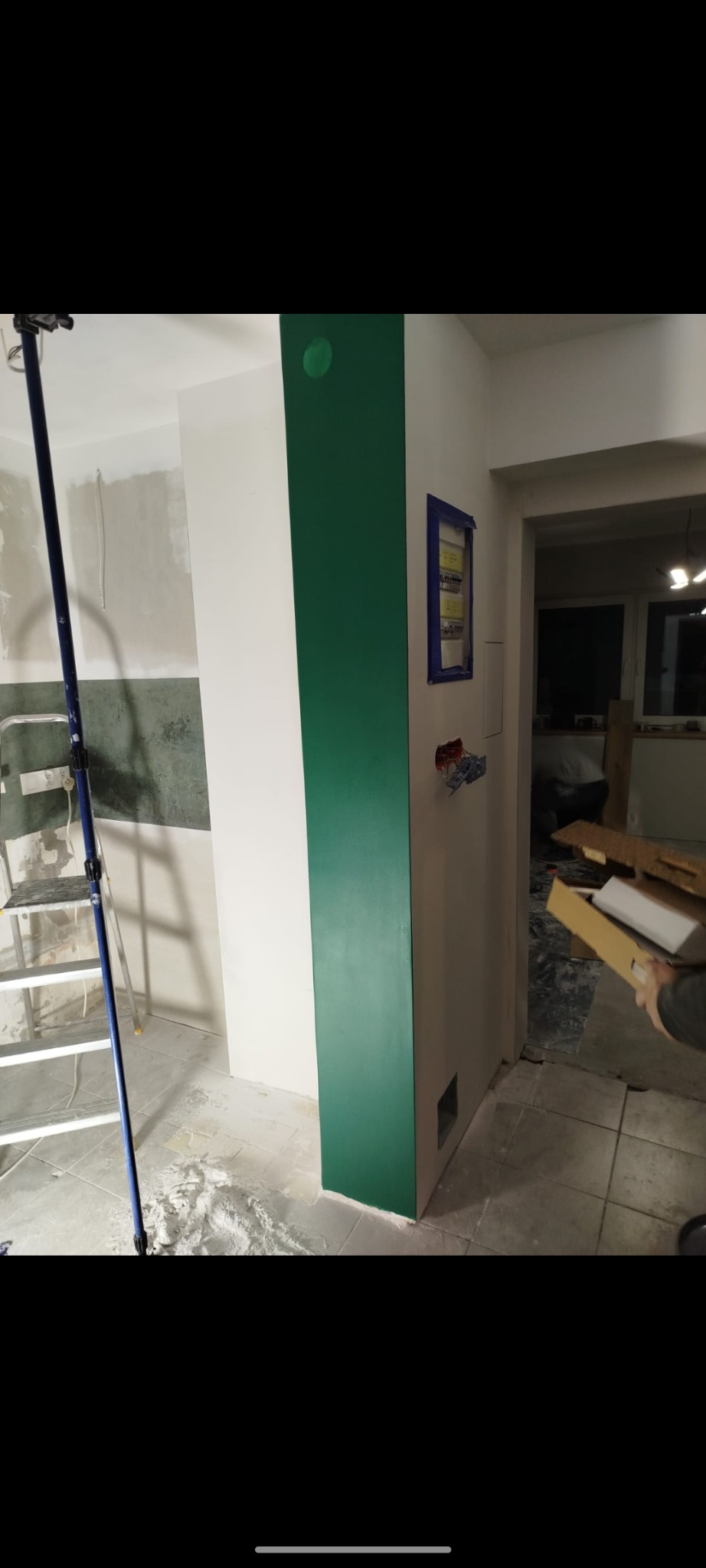 Wnętrze w trakcie prac: ściana z fragmentem pomalowanym na intensywny zielony kolor, widoczna drabina malarska, niezabezpieczone przewody elektryczne i rozdzielnia, ślady gładzi na podłodze.