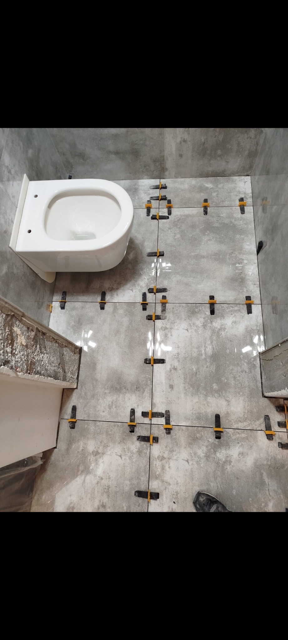 Układanie szarych płytek podłogowych w łazience z zamontowaną białą miską WC, widoczne czarne krzyżyki dystansowe i żółte kliny poziomujące.