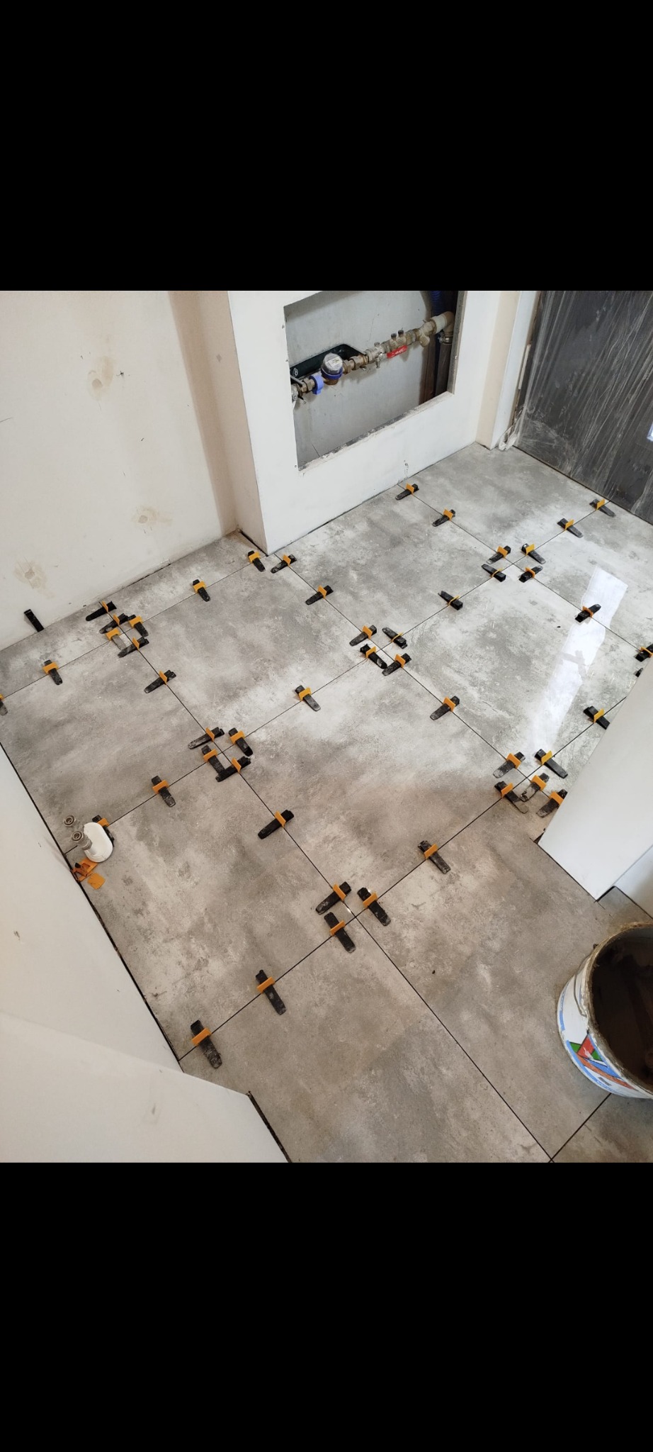 Szare płytki podłogowe w trakcie układania, z użyciem klipsów poziomujących, widoczna puszka z instalacją wodną w ścianie i wiadro z zaprawą.