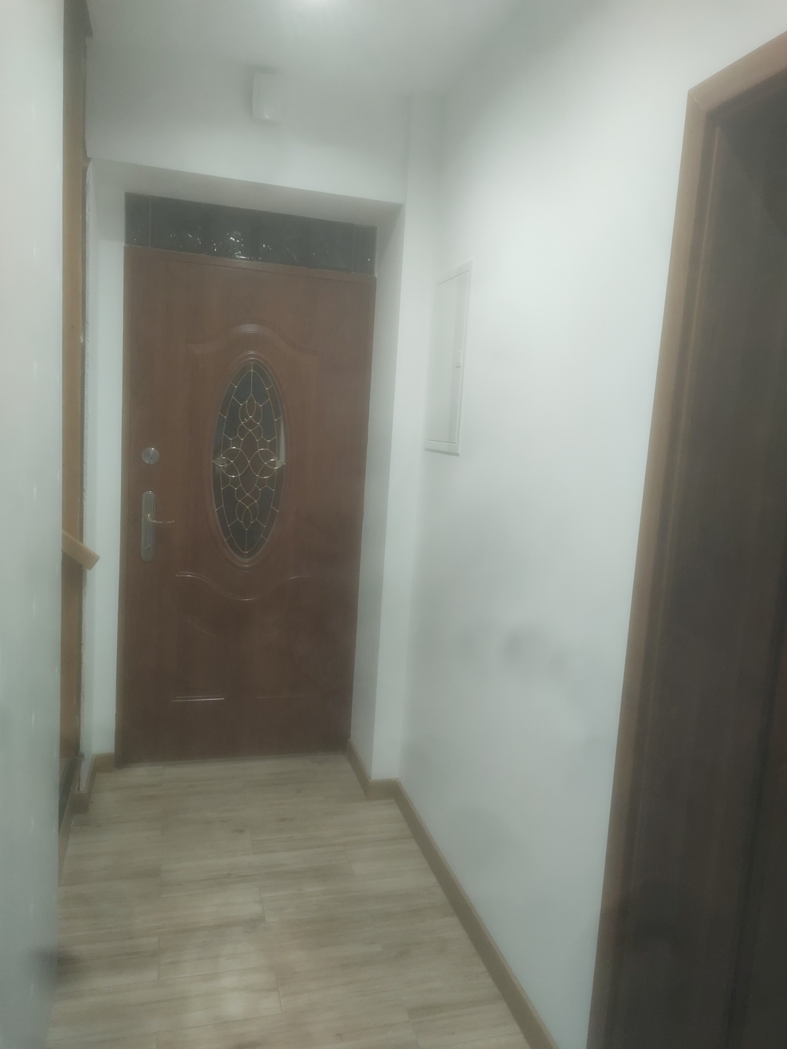 Korytarz po remoncie z brązowymi panelami podłogowymi imitującymi drewno oraz brązowymi drzwiami wejściowymi z szybą z ozdobnym wzorem, ściany pomalowane na biało.