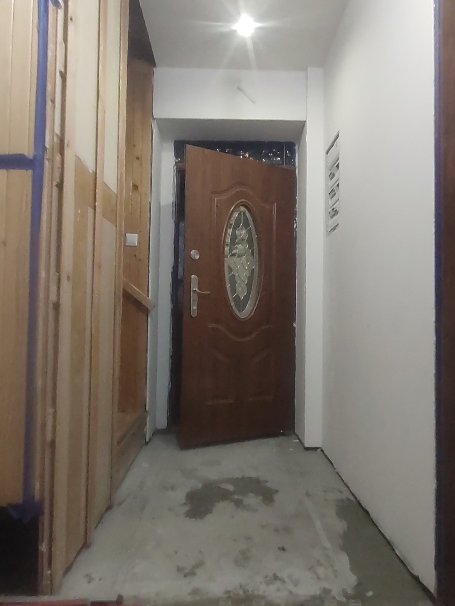 Korytarz w trakcie remontu: widoczne drewniane panele ścienne w trakcie montażu, otwarta brązowa brama wejściowa z ozdobnym szkłem, surowa podłoga betonowa i białe ściany.
