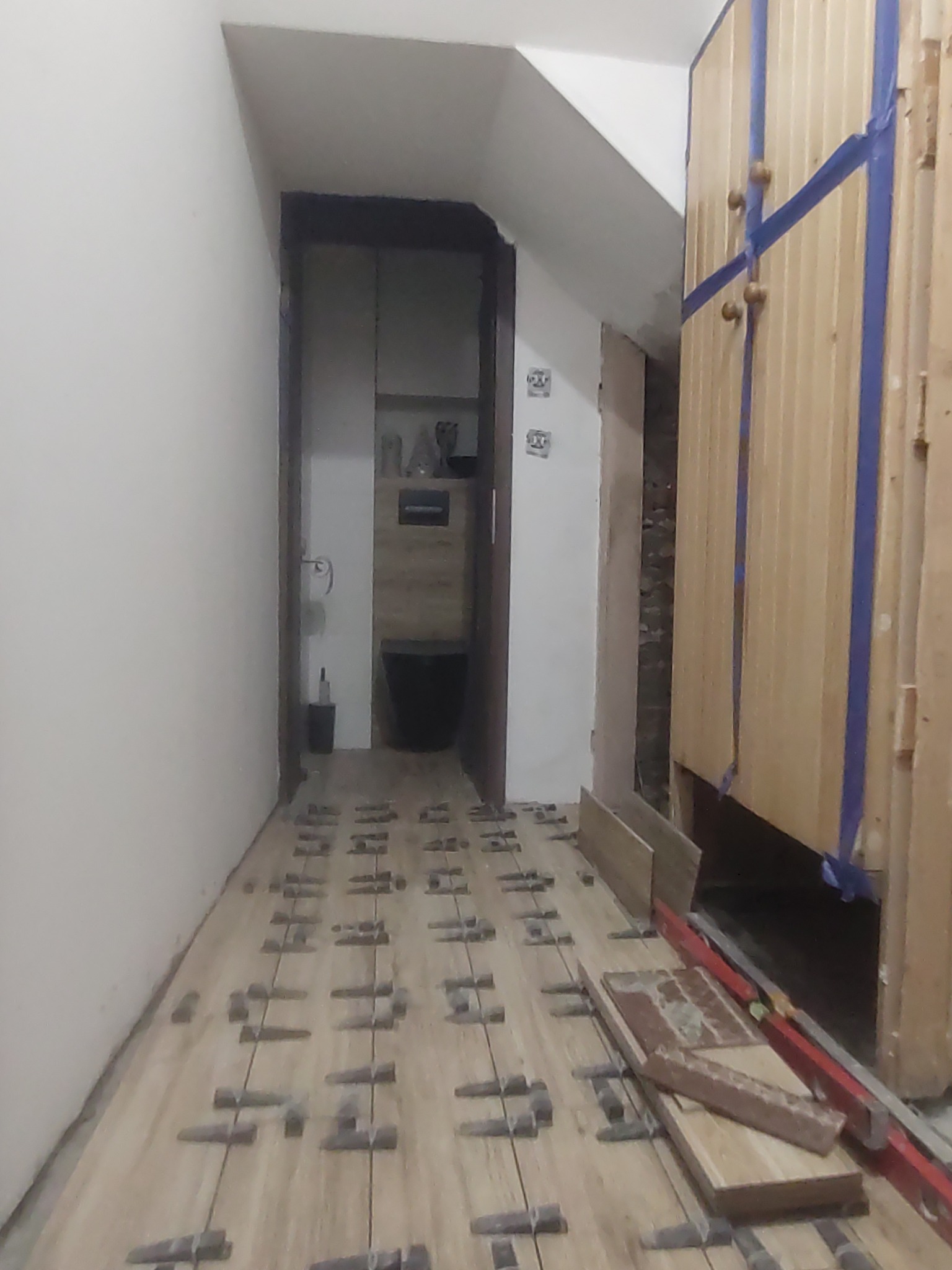 Korytarz w trakcie kładzenia podłogi z płytek imitujących drewno, widoczne kliny dystansowe, płytki przygotowane do ułożenia, drzwi do łazienki w tle.