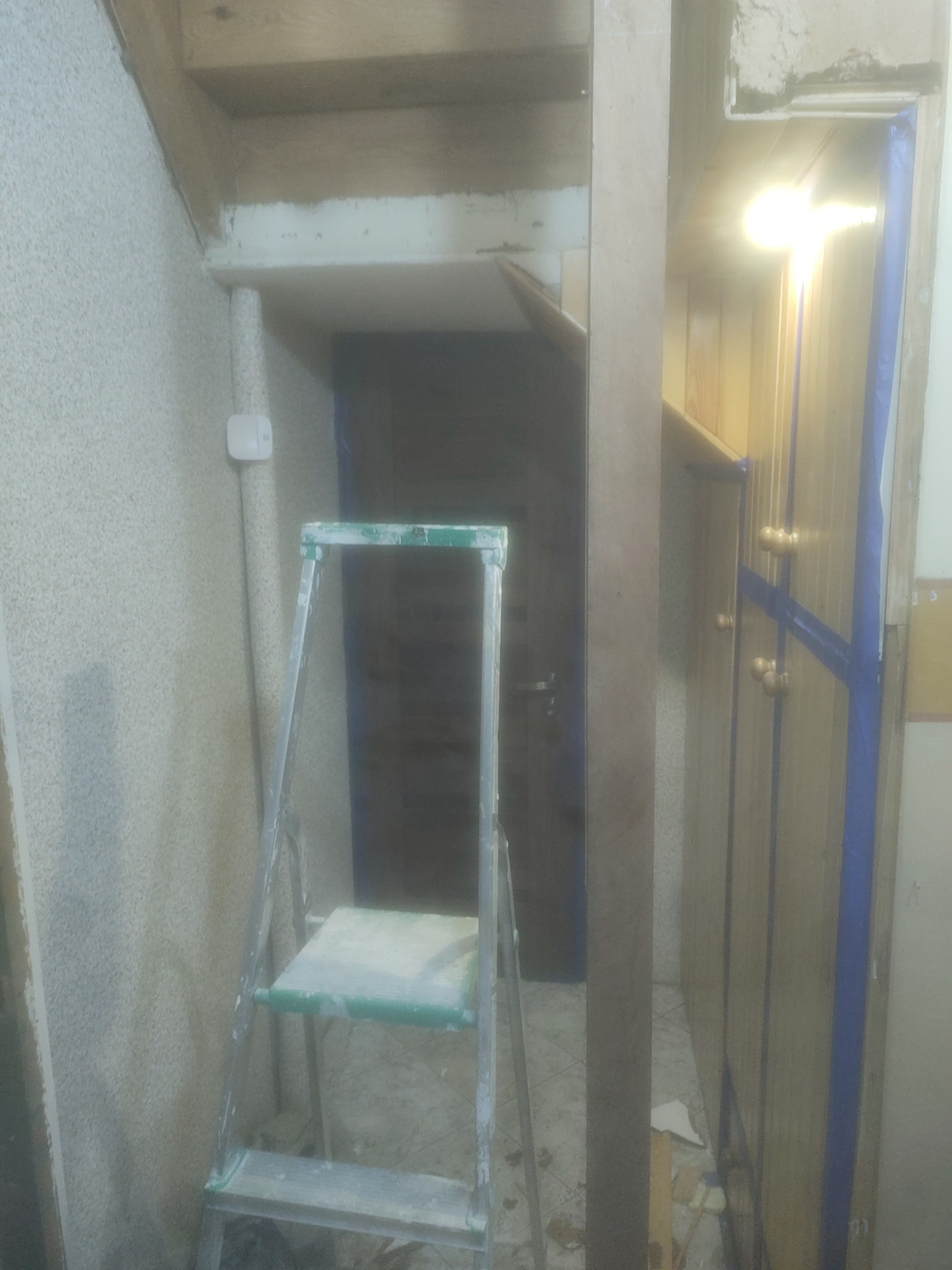 Wnętrze w trakcie prac: drabina aluminiowa oparta o ścianę z grubym tynkiem strukturalnym, zabezpieczone folią drzwi szafy wnękowej z drewnianymi uchwytami, widoczne ślady po gładzi na suficie.