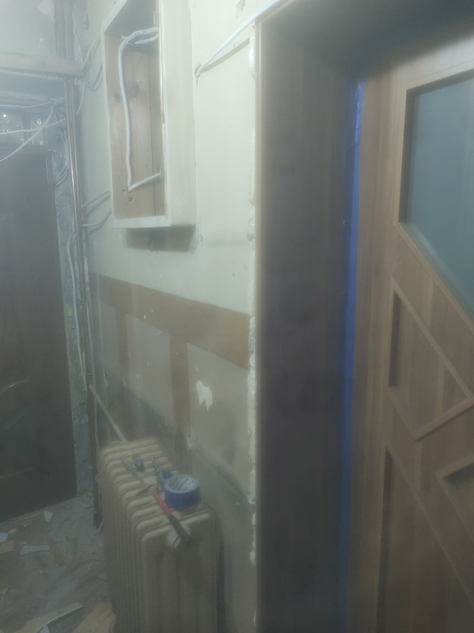 Korytarz w trakcie prac przygotowawczych, widoczne zabezpieczone taśmą malarską ościeżnice, grzejnik z narzędziami i niedokończone obudowy instalacji.