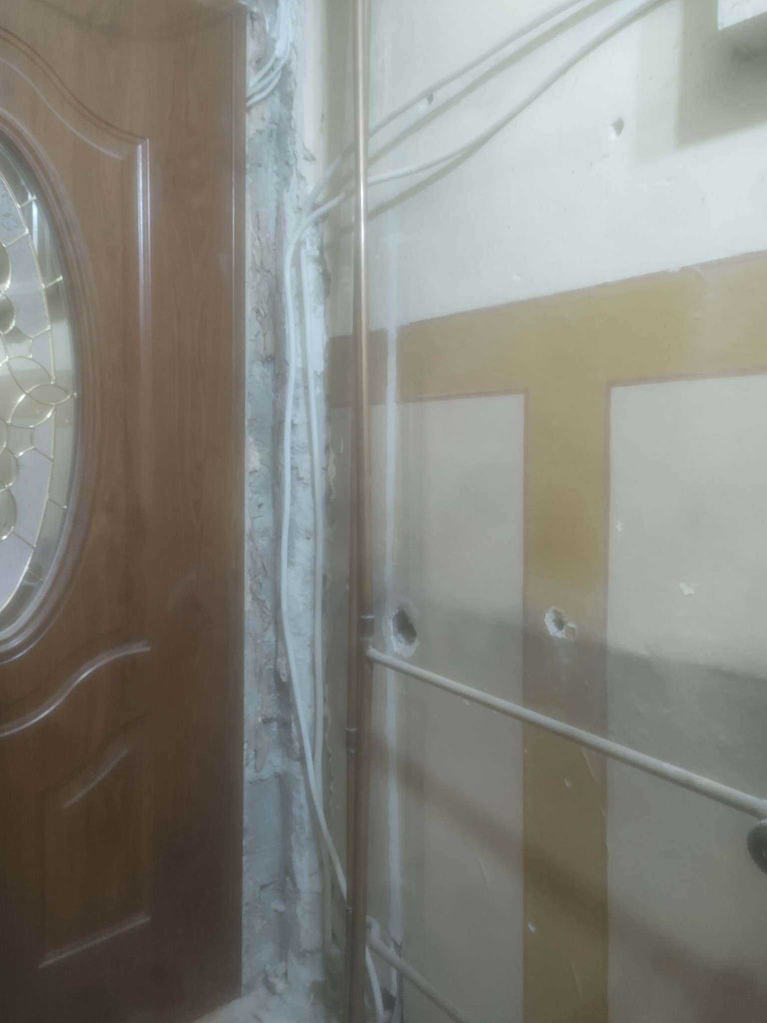 Fragment ściany w trakcie prac remontowych z widocznymi przewodami, rurami i uszkodzonym tynkiem obok drewnianych drzwi z szybą.