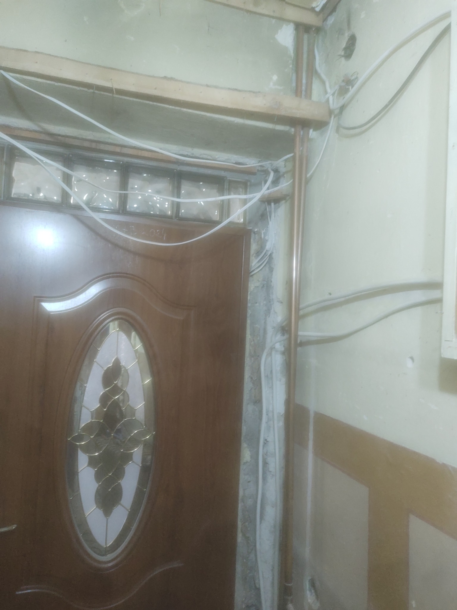 Remont wejścia do mieszkania w Bolszewie: widoczne przewody elektryczne, rury miedziane, surowy mur i częściowo zamontowane szklane luksfery nad drzwiami z owalnym przeszkleniem.