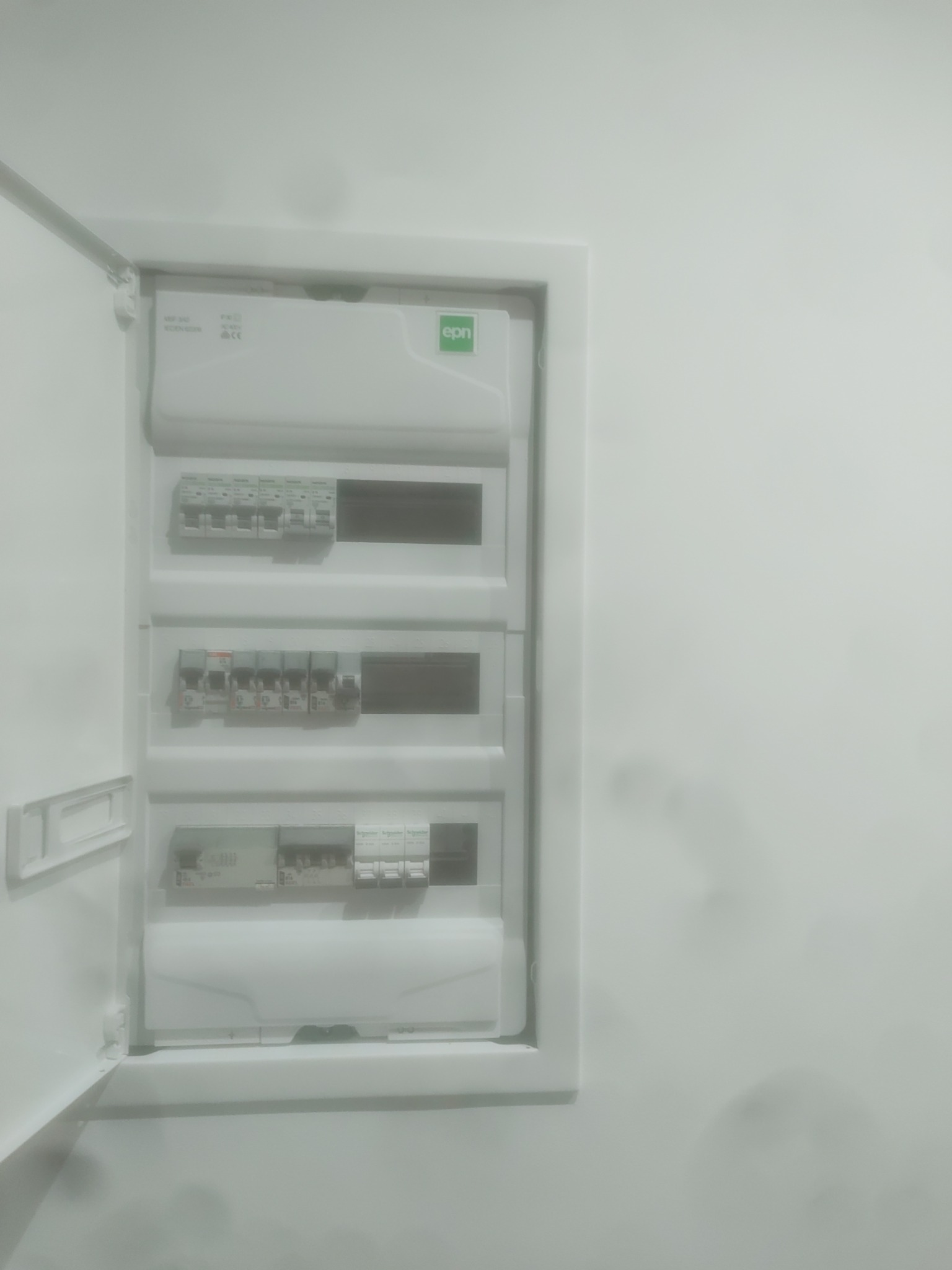 Otwarta, biała skrzynka elektryczna z bezpiecznikami na białej ścianie, widoczne elementy instalacji elektrycznej.