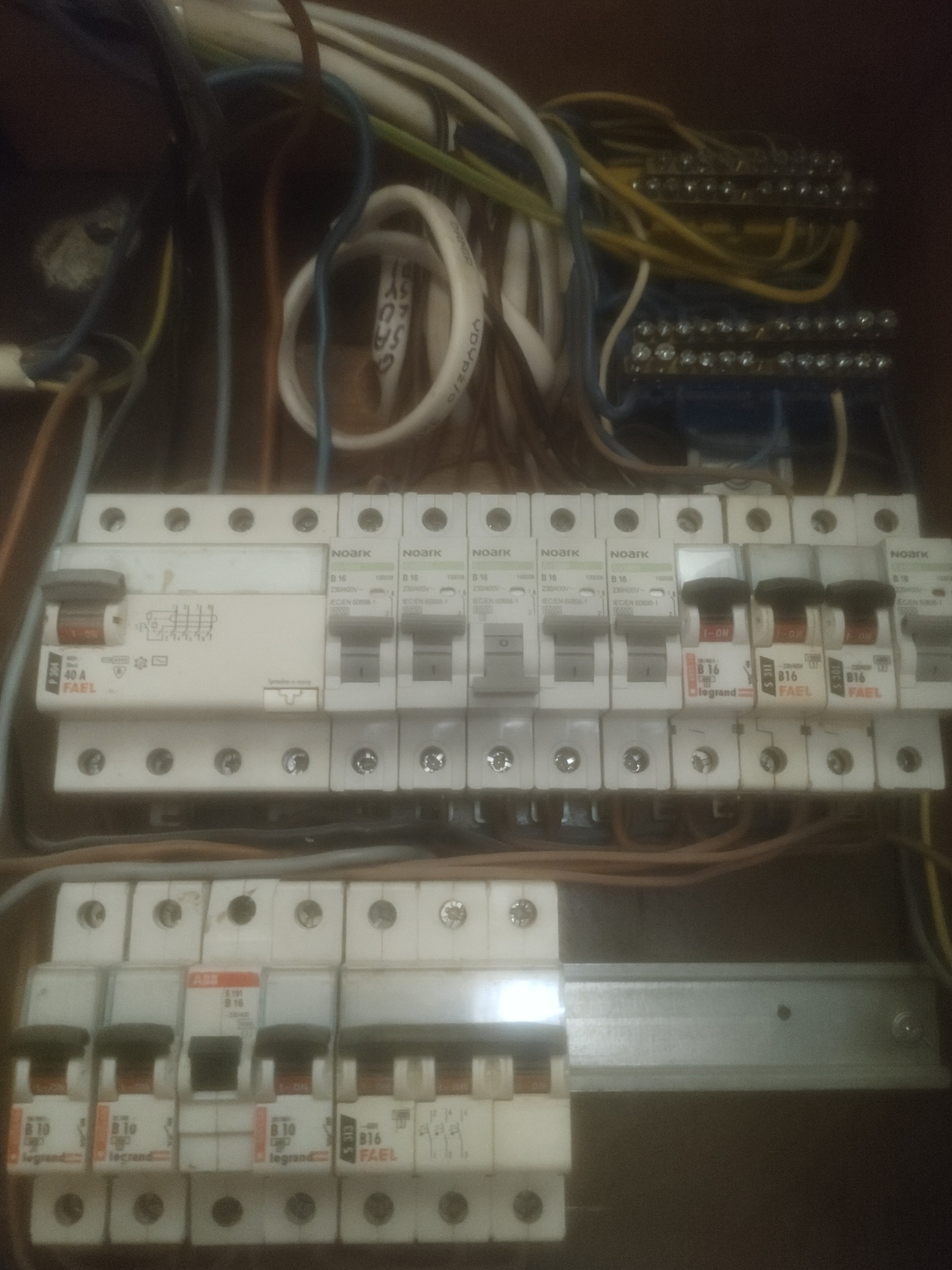 Skrzynka elektryczna z bezpiecznikami różnych marek (Noark, Legrand, Fael, ABB) i różnymi wartościami prądu, widoczne przewody i listwy zaciskowe, wykonane z bliska w słabym oświetleniu.