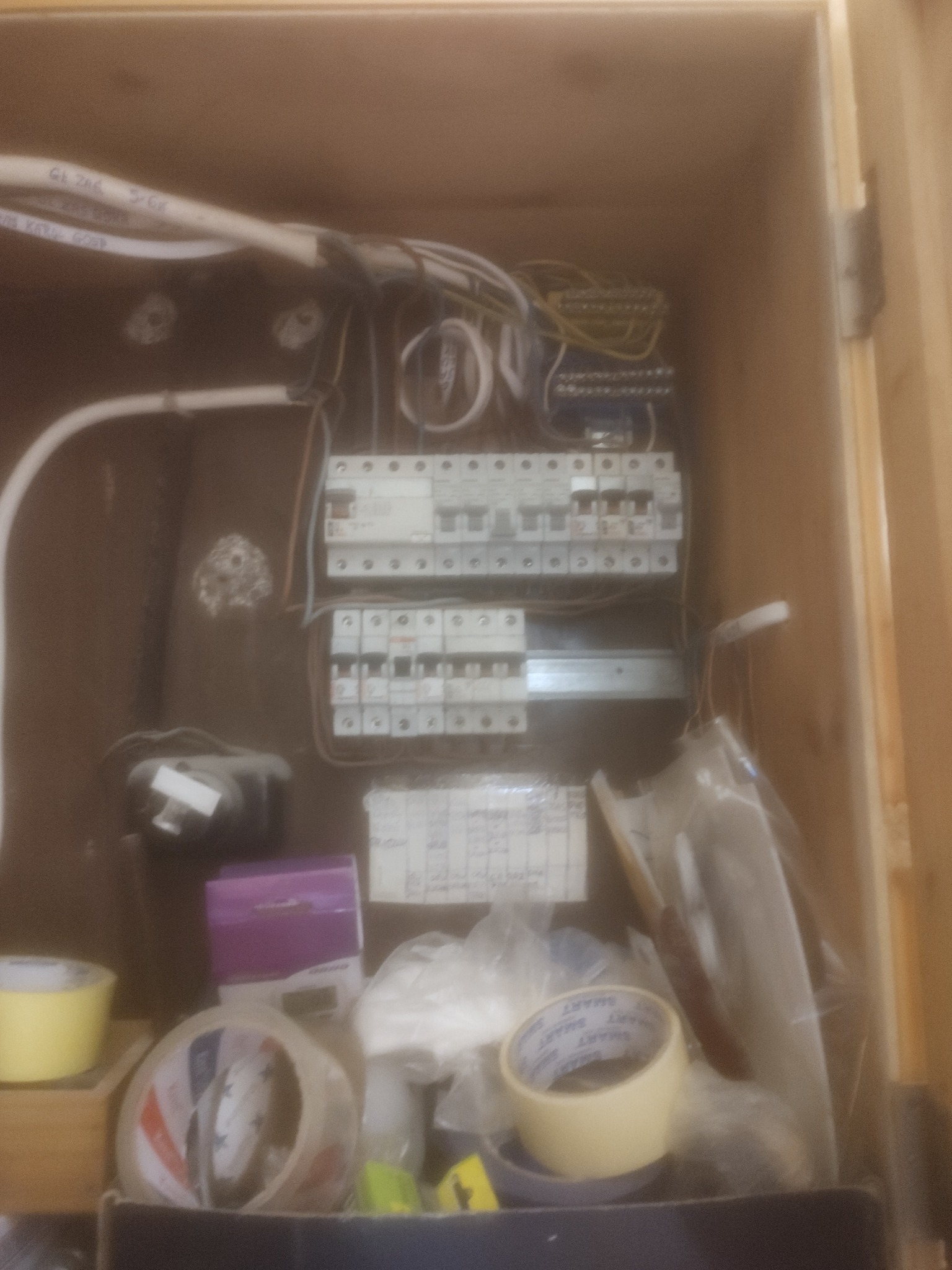 Otwarta skrzynka elektryczna z widocznymi bezpiecznikami, przewodami i akcesoriami montażowymi, w tym taśmami izolacyjnymi i pudełkiem po akcesoriach elektrycznych.