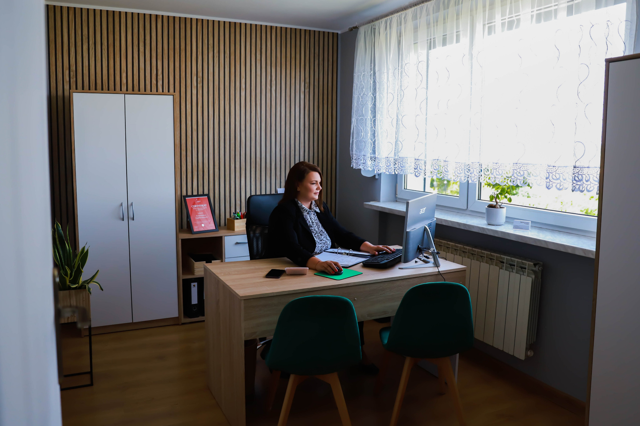 Pracownica biura przy biurku z komputerem, w tle regał z certyfikatem, drewniana ściana i okno z firanką. Jasne, przestronne wnętrze biura.