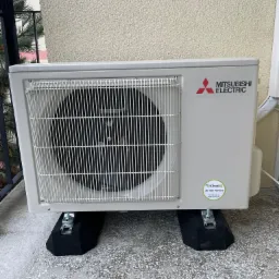 Zewnętrzna jednostka klimatyzacji Mitsubishi Electric zamontowana na czarnych, tłumiących drgania podstawach, z widocznym logo producenta i naklejką firmy instalacyjnej, na tle beżowej ściany budynku.