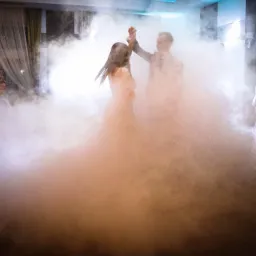 Para młoda tańczy pierwszy taniec w gęstej mgle podczas przyjęcia weselnego, goście obserwują.