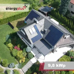 Widok z drona na dom z panelami słonecznymi na dachu, zadbany ogród z różnorodną roślinnością, logo firmy fotowoltaicznej w lewym górnym rogu i oznaczenie mocy instalacji 9,8 kWp w prawym dolnym rogu.