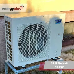 Biała jednostka zewnętrzna klimatyzacji GREE Versati 10 kW, zamontowana na metalowej konstrukcji, widoczna na tle ściany budynku i okna piwnicznego.