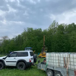 Żółta wiertnica do studni na trawiastym terenie, biały samochód terenowy z przyczepą cysterną w tle lasu i pochmurnego nieba.