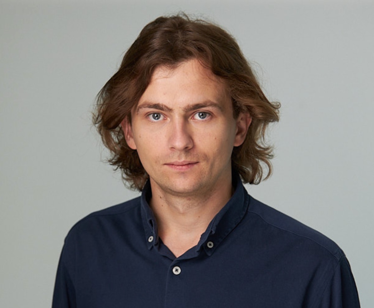 Portret mężczyzny z brązowymi włosami i niebieskimi oczami, ubrany w granatową koszulę na tle szarego tła.