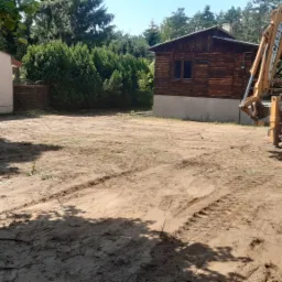 Niwelacja terenu pod budowę z widocznym traktorem Case 580 Super F i budynkami w tle, ślady opon na wyrównanym gruncie.