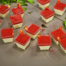 Deser w przezroczystych kwadratowych pojemnikach: kremowa baza i czerwona galaretka truskawkowa, udekorowany kawałkiem truskawki, rozstawiony na szarym tle z zielonymi liśćmi.