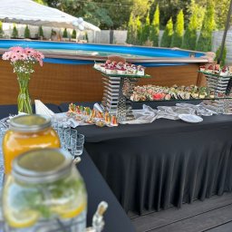 Catering Malwa - Stół z czarnym obrusem, na którym znajdują się przekąski, sushi, desery, napoje w słojach z kranikami, szklanki, wazon z różowymi kwiatami, w tle basen i zielone drzewa.