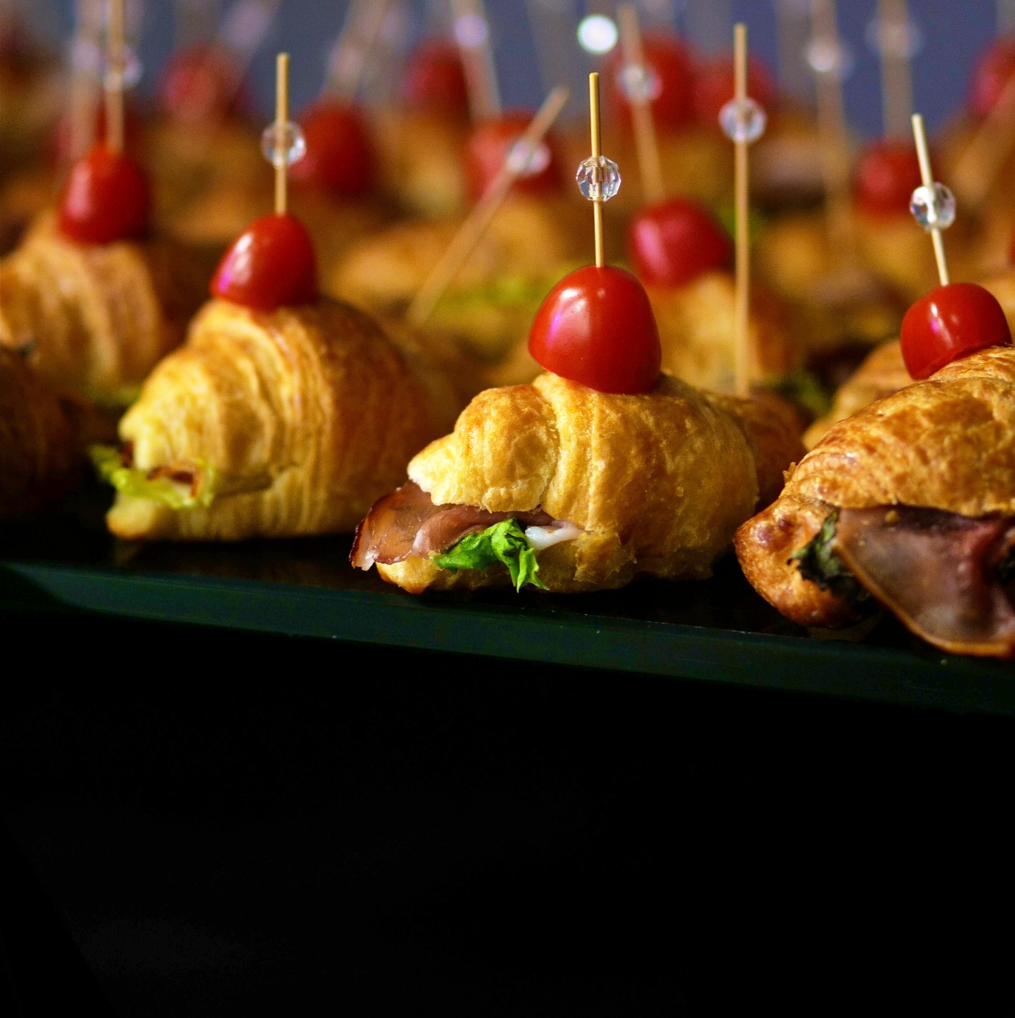 Rząd mini croissantów z szynką, sałatą i pomidorkiem cherry na ozdobnym patyczku, ułożonych na szklanej tacy.