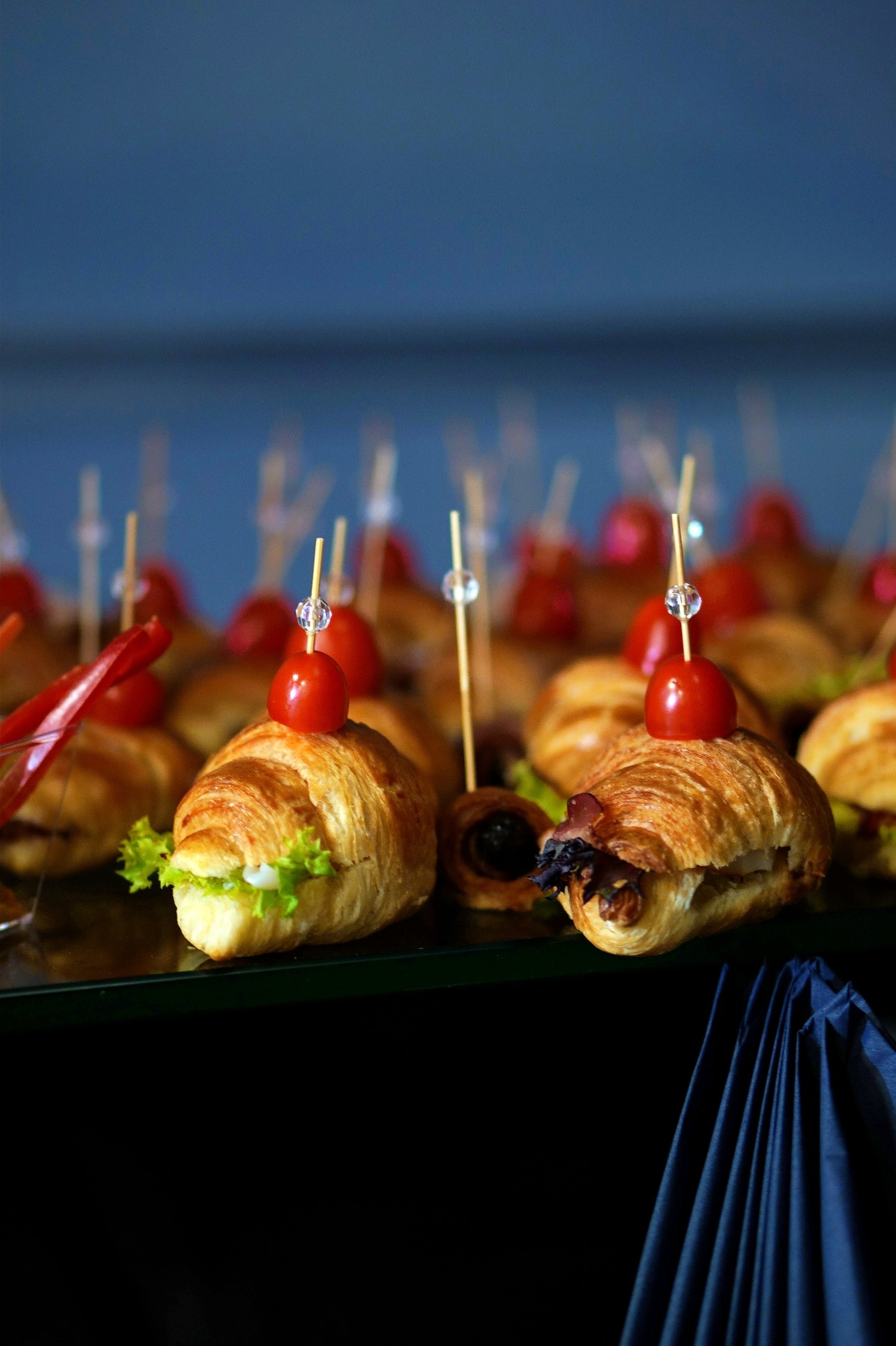 Kruche rogaliki z nadzieniem i pomidorkiem cherry na wykałaczce, ułożone na szklanym stole z błękitnym tłem, widoczne detale nadzienia i dekoracyjne elementy wykałaczek.