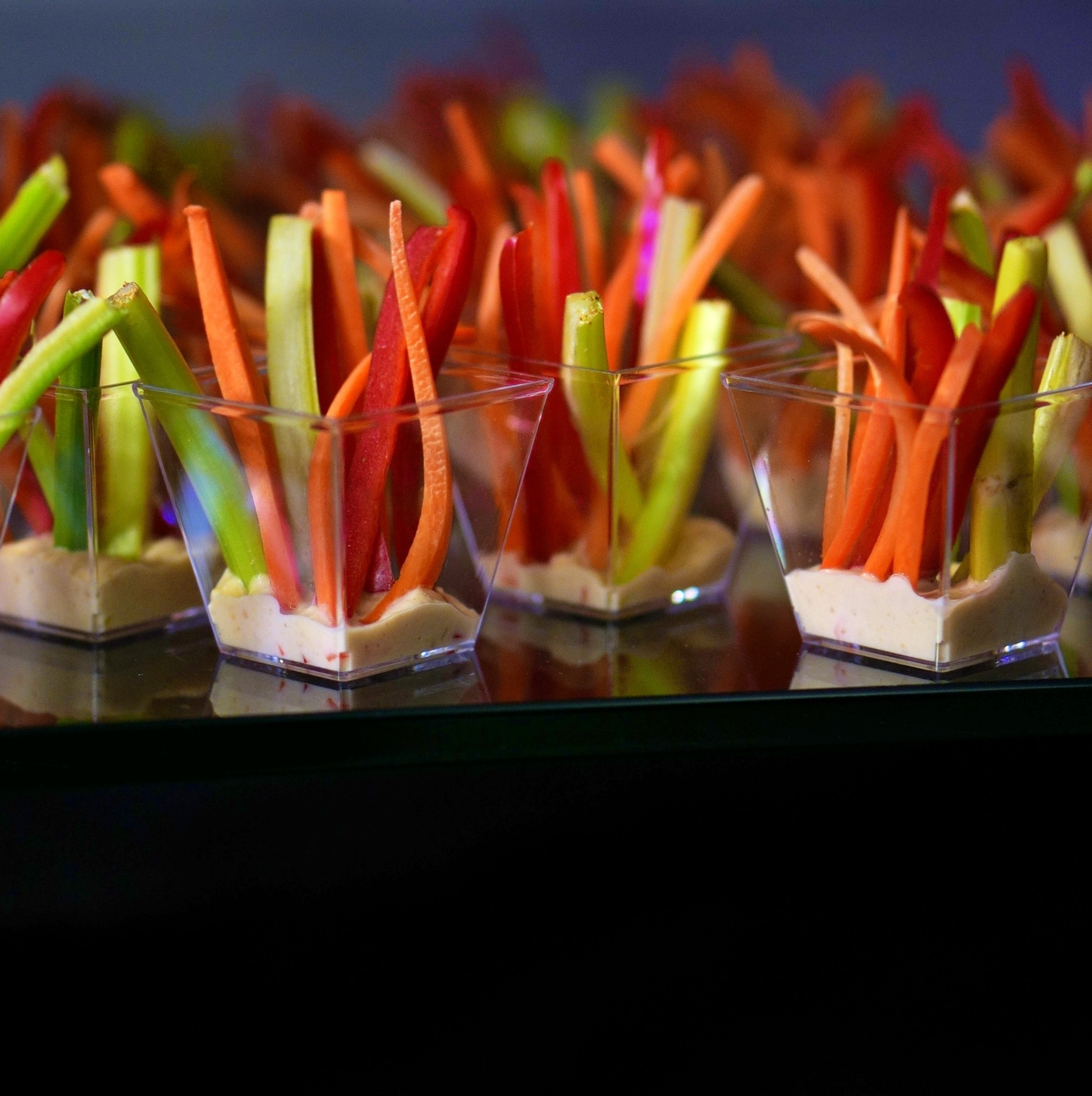 Elegancko podane warzywa (seler naciowy, marchew, papryka) w słupkach, umieszczone w małych, przezroczystych naczyniach z jasnym dipem, ustawione rzędami na ciemnym, błyszczącym podłożu.