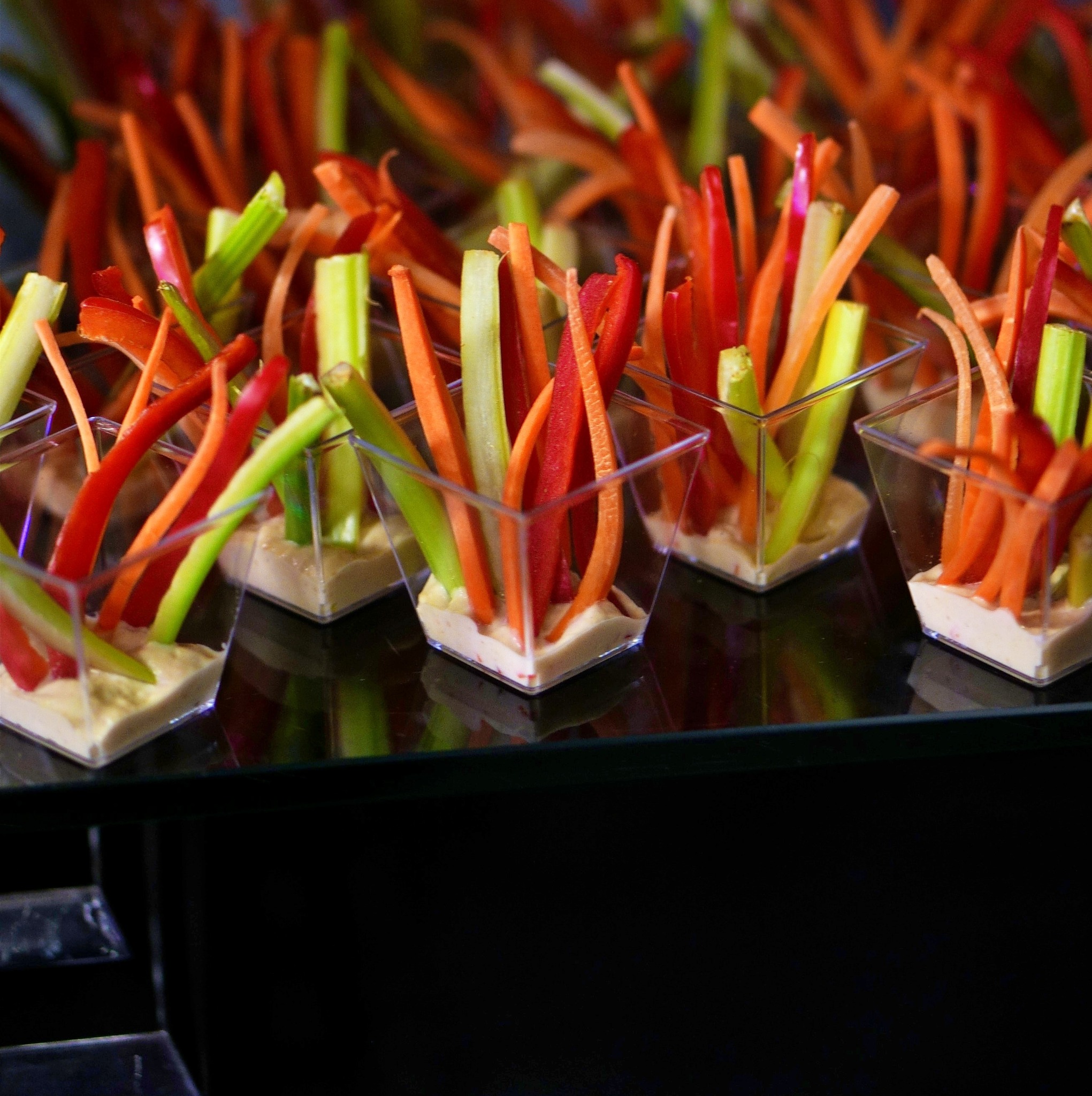 Szklane naczynia z dipem i warzywnymi słupkami: marchew, seler naciowy i czerwona papryka, ułożone na czarnym, błyszczącym stole.
