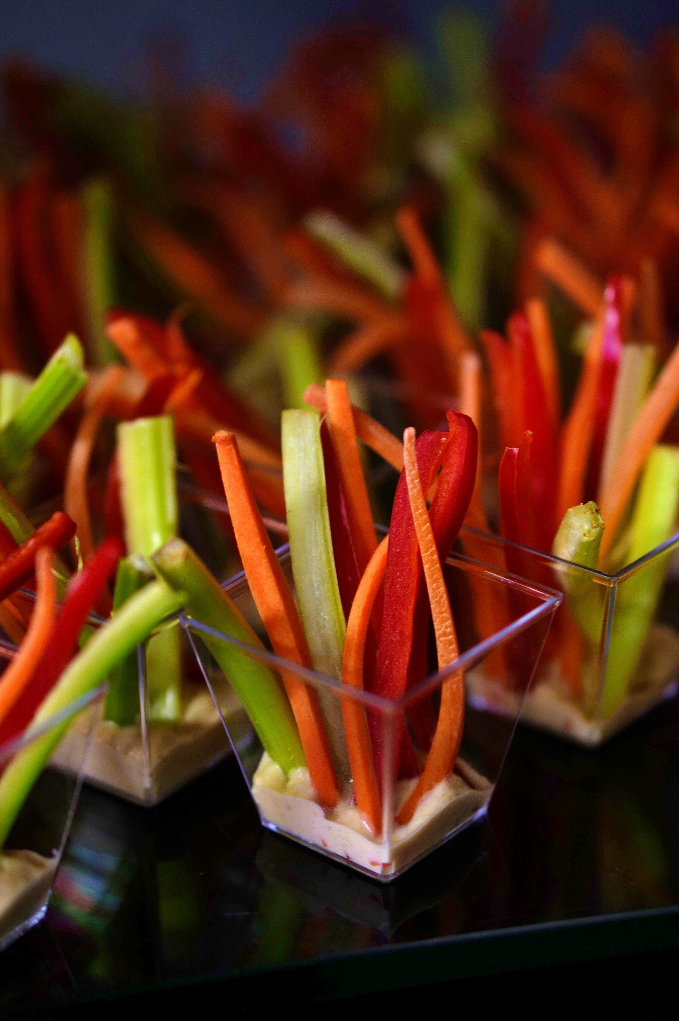 Miniaturowe porcje warzyw (papryka, seler naciowy, marchew) pokrojonych w słupki, zanurzonych w kremowym dipie, podane w przezroczystych, kwadratowych naczyniach na ciemnej, błyszczącej powierzchni.