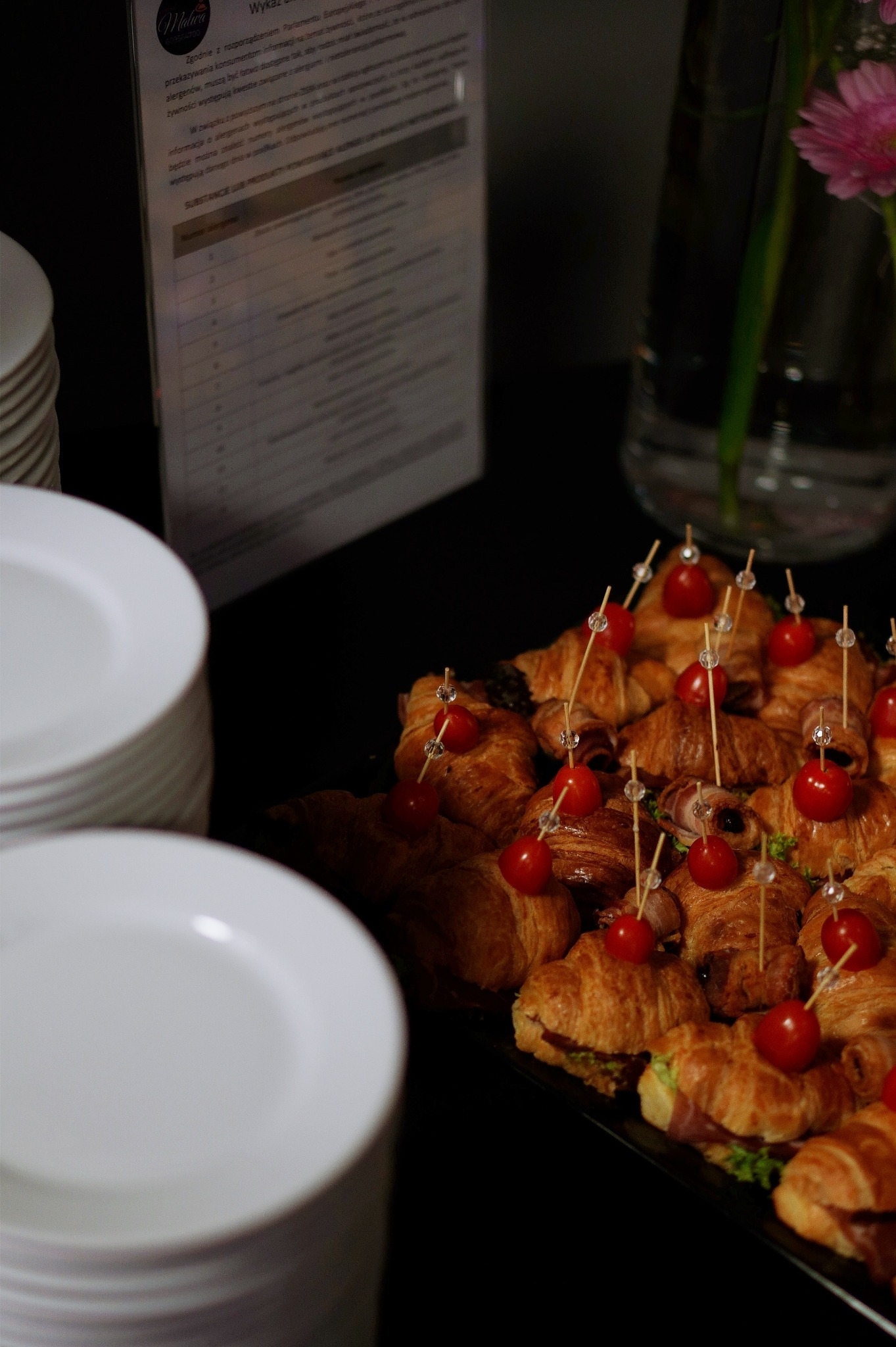 Kruche rogaliki z szynką i sałatą, udekorowane pomidorkami cherry na wykałaczkach z ozdobnymi końcówkami, ułożone na czarnym talerzu obok stosu białych talerzy, w tle karta menu i wazon z kwiatami.