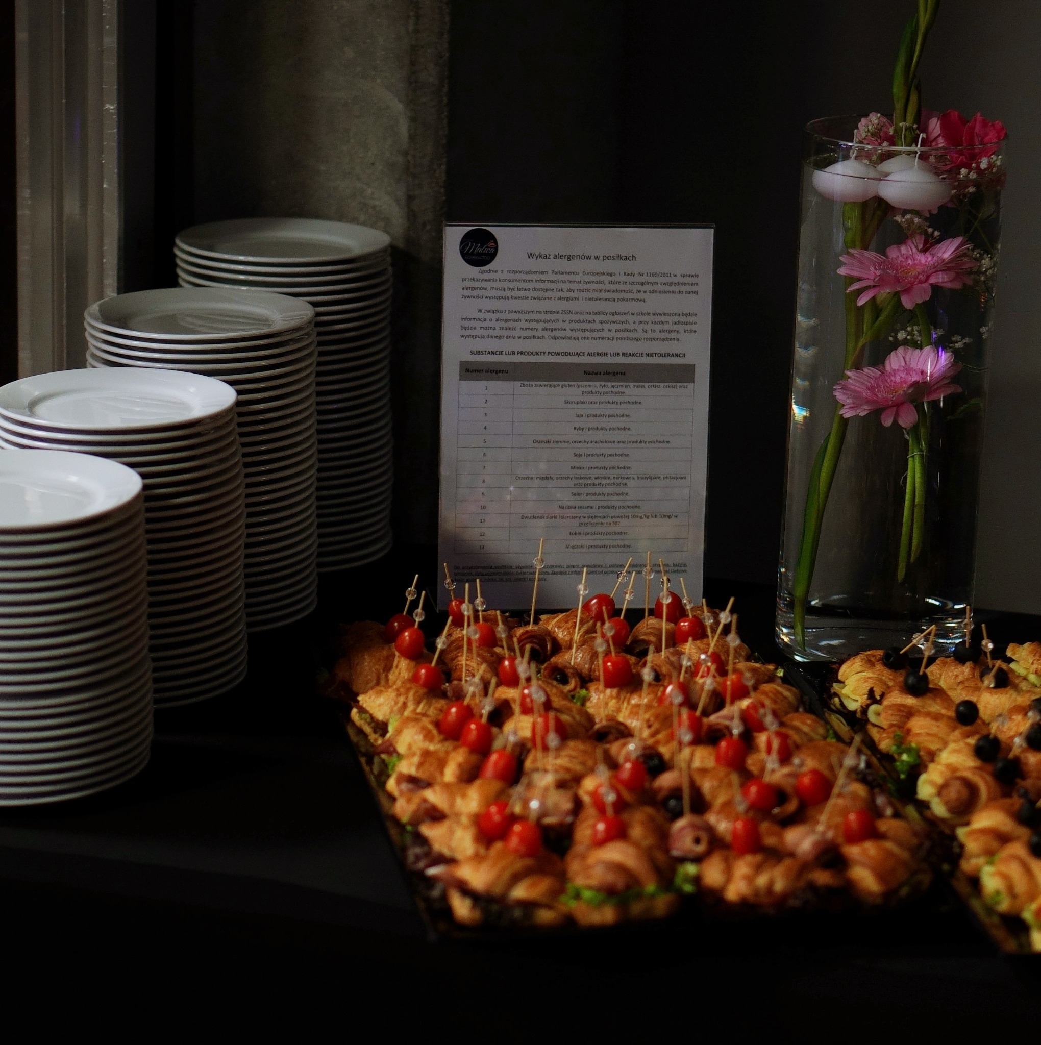Ułożone na czarnym stole przekąski na catering: mini croissanty z pomidorkami cherry na wykałaczkach, obok stosy białych talerzy, w tle wazon z różowymi kwiatami i tabliczka informująca...