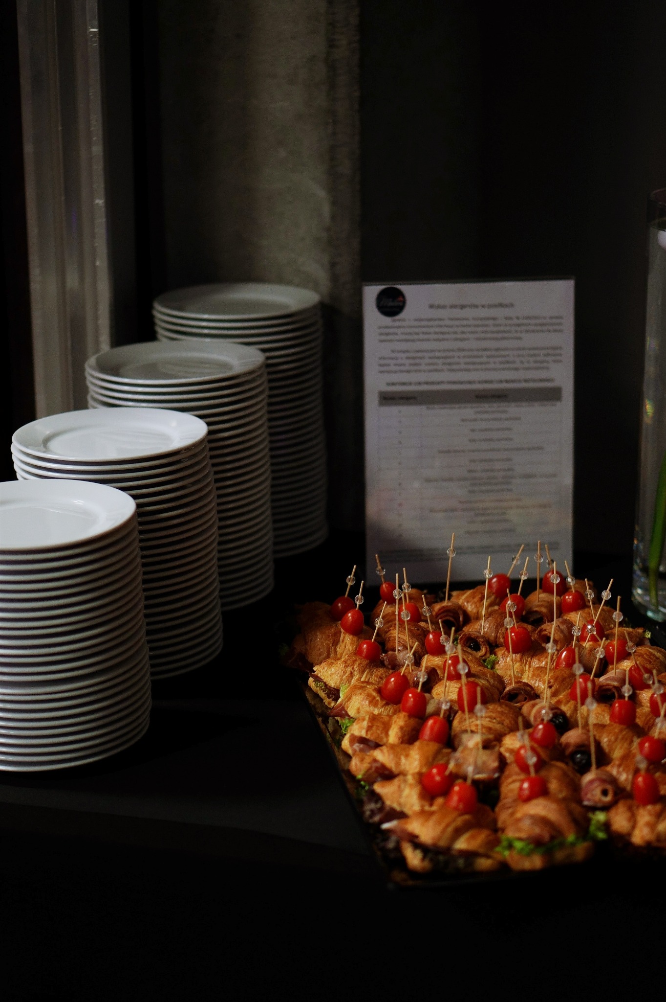 Kruche rogaliki z szynką i pomidorkami koktajlowymi na czarnej tacy, obok stosy białych talerzy i menu w ramce, ustawione na ciemnym stole.