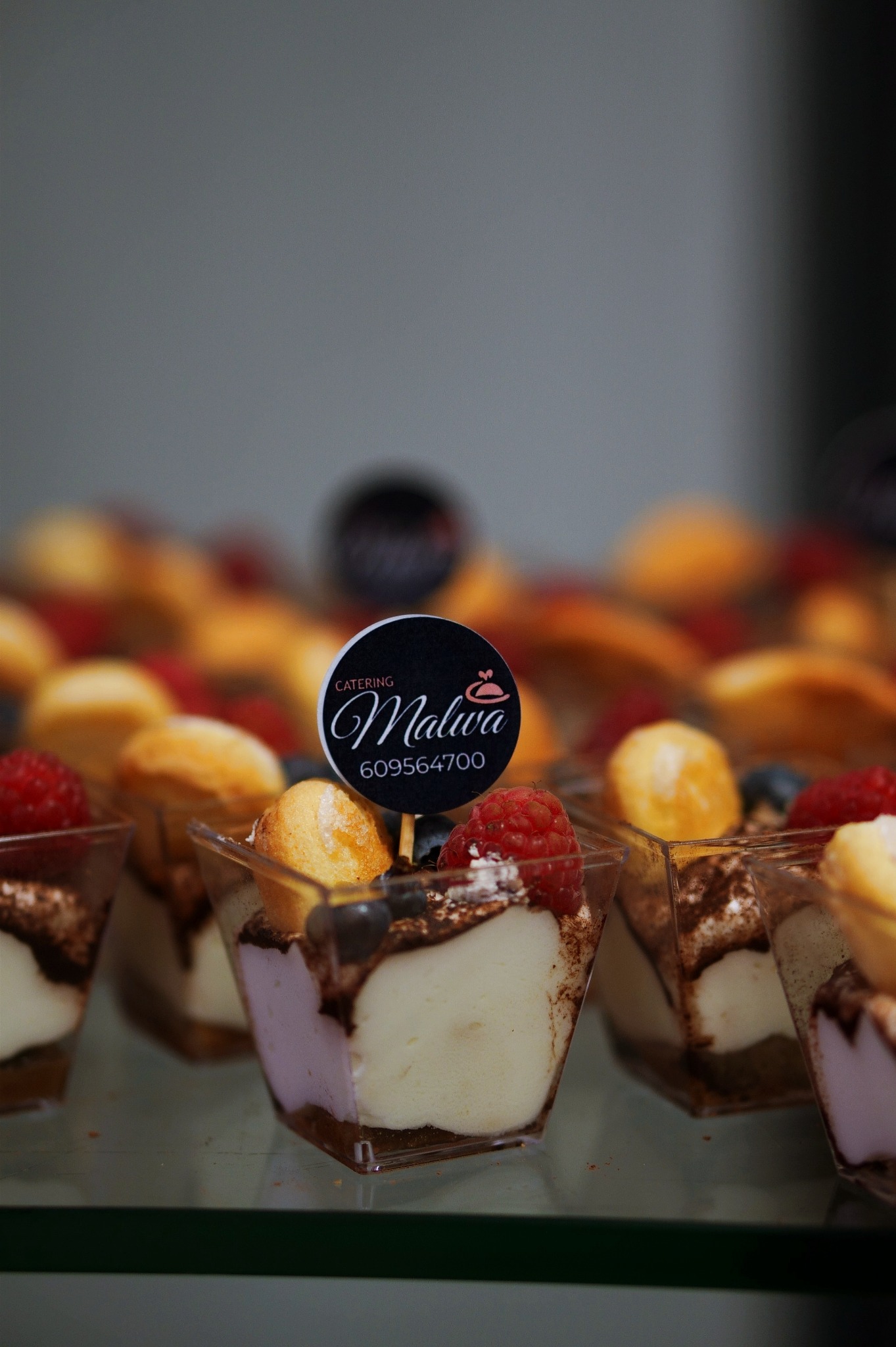 Mini deserki w plastikowych pucharkach, z kremem, owocami i ciasteczkiem, ozdobione logo firmy cateringowej na piku.