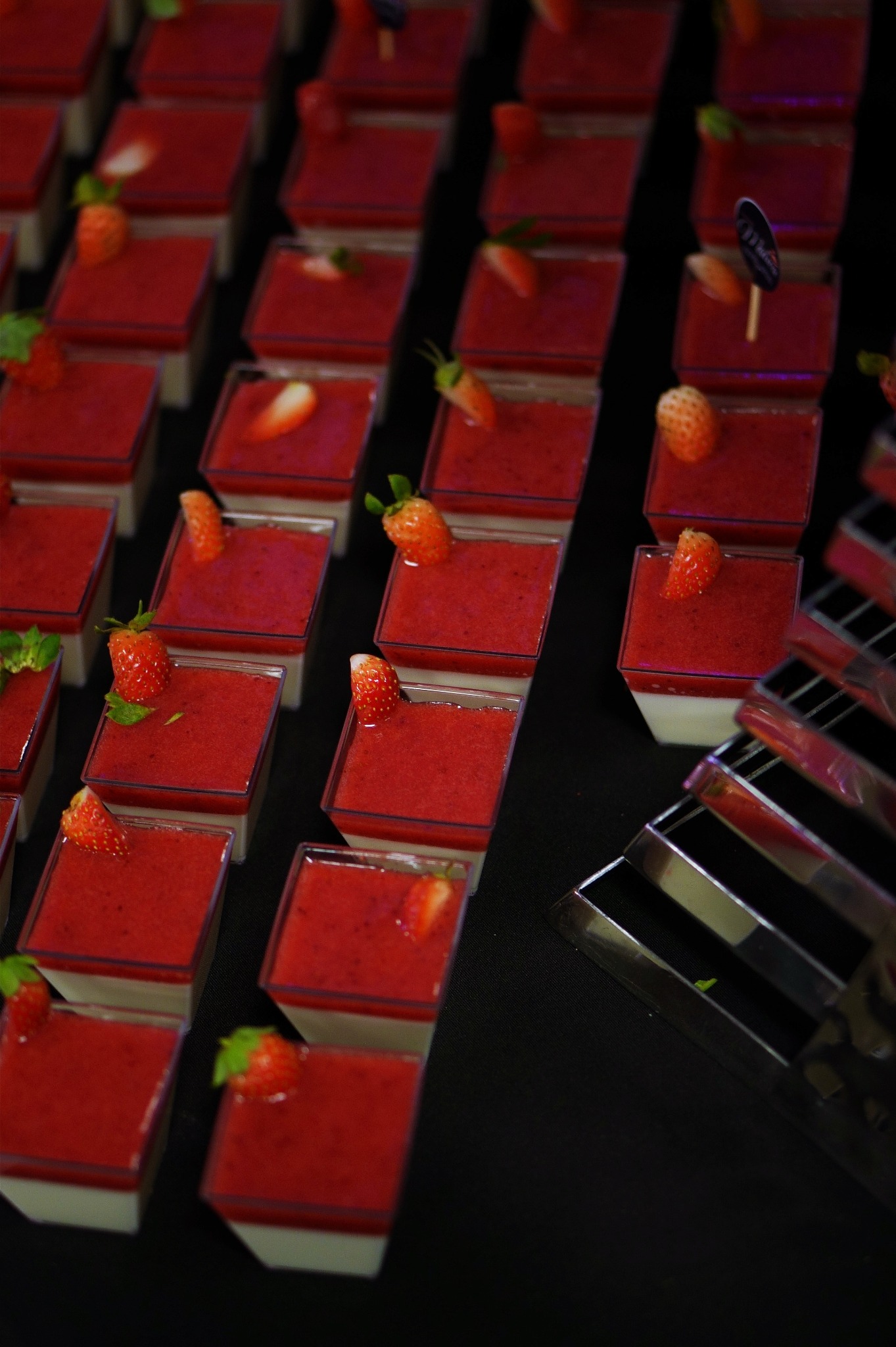 Rządki deserów w plastikowych pojemnikach, każdy z warstwą białej pianki i czerwoną galaretką truskawkową, udekorowane świeżymi truskawkami i listkami mięty, ustawione na czarnym obrusie, widoczny...