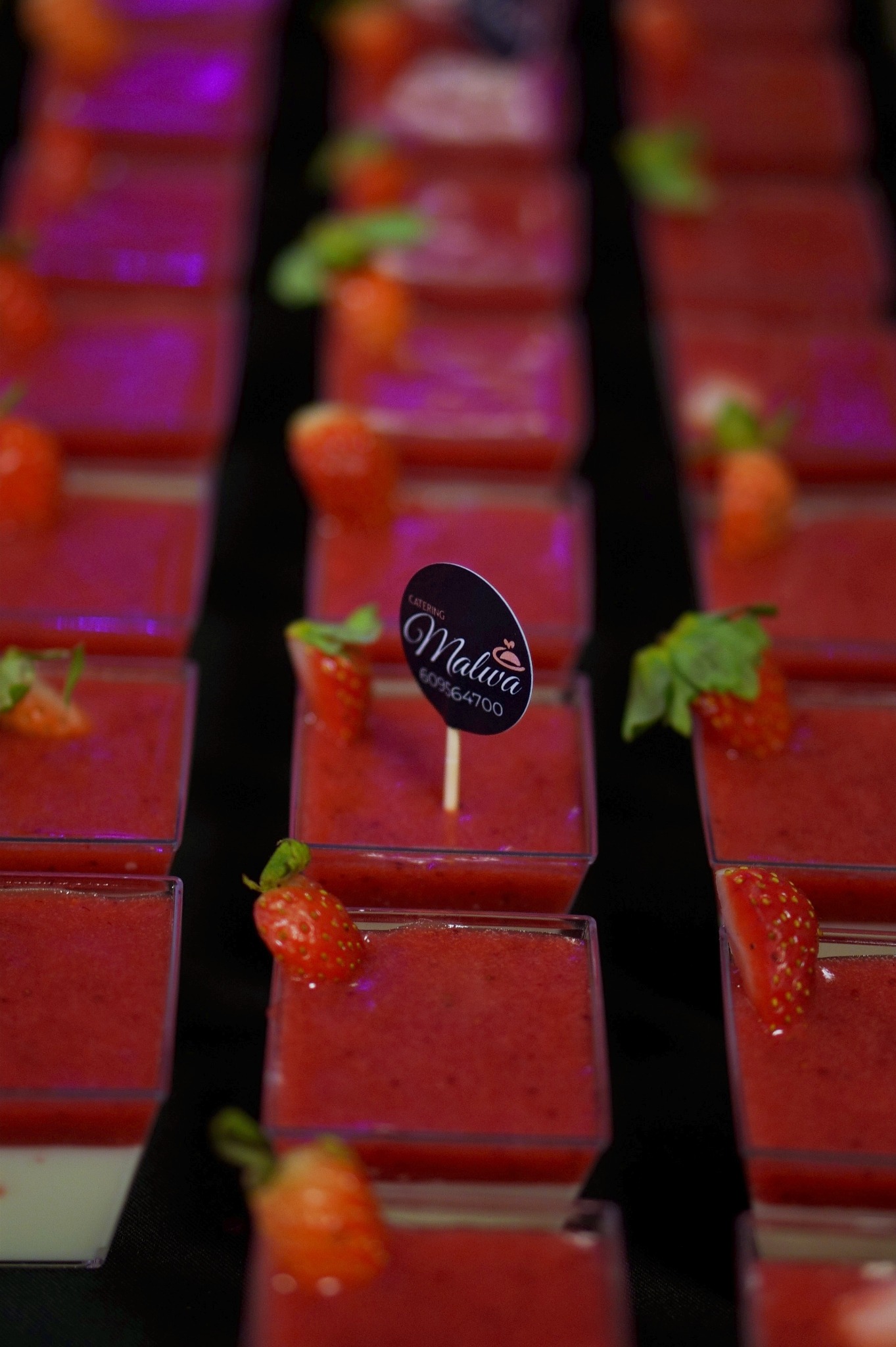 Rzędy deserów truskawkowych w przezroczystych pojemnikach, udekorowanych połówkami truskawek i logiem firmy cateringowej na patyczku, podświetlone fioletowym światłem.