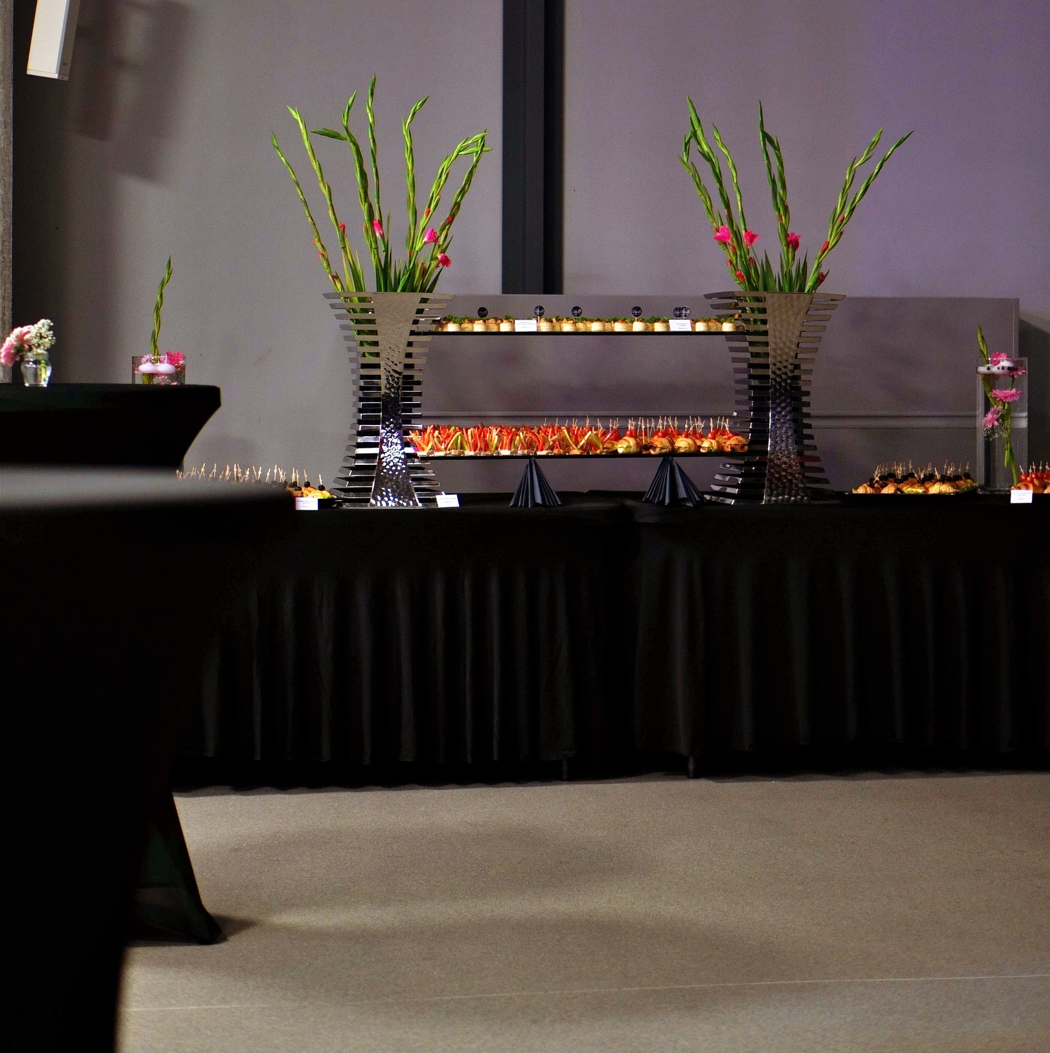 Elegancki catering: stół z czarnym obrusem, metalowa konstrukcja z przekąskami na szklanych półkach, dekoracje z wysokich kwiatów i małe bukiety w szklanych wazonach.