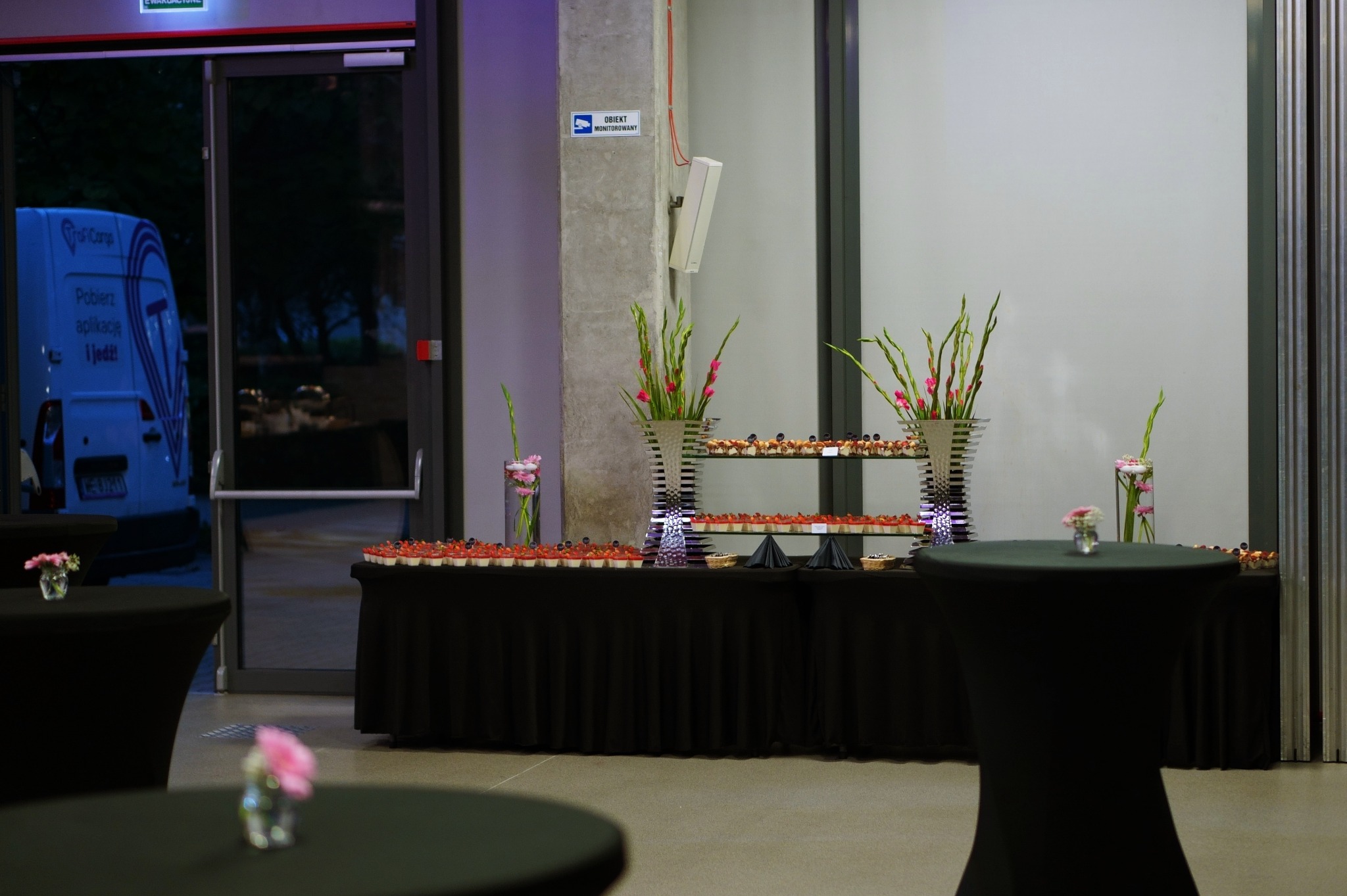 Stół z czarnym obrusem, na którym znajdują się desery, kwiaty w wazonach i dwupoziomowy srebrny stojak z przekąskami, w tle drzwi ewakuacyjne i betonowy filar z kamerą monitoringu.