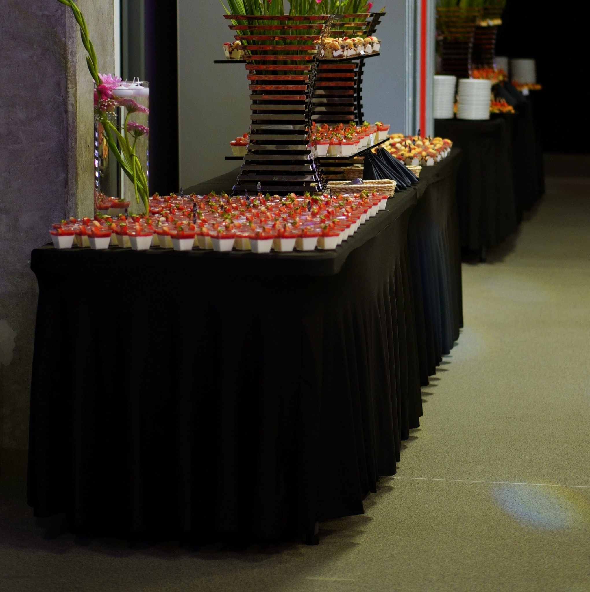 Długi stół z czarnym obrusem prezentuje rzędy deserów w małych pojemnikach z czerwonymi owocami na wierzchu, dekoracje kwiatowe w wazonach i nowoczesne, wielopoziomowe stojaki z przekąskami...