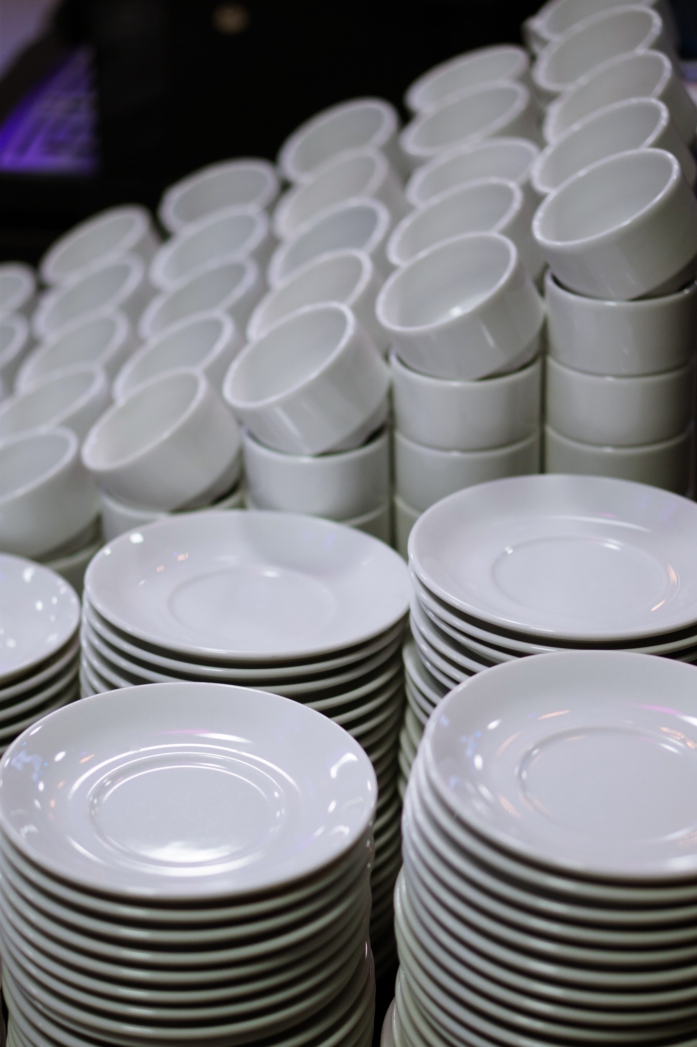Stosy białych, okrągłych talerzy i misek ułożone w rzędach, przygotowane do serwowania, oświetlone z góry, widoczne delikatne refleksy światła.