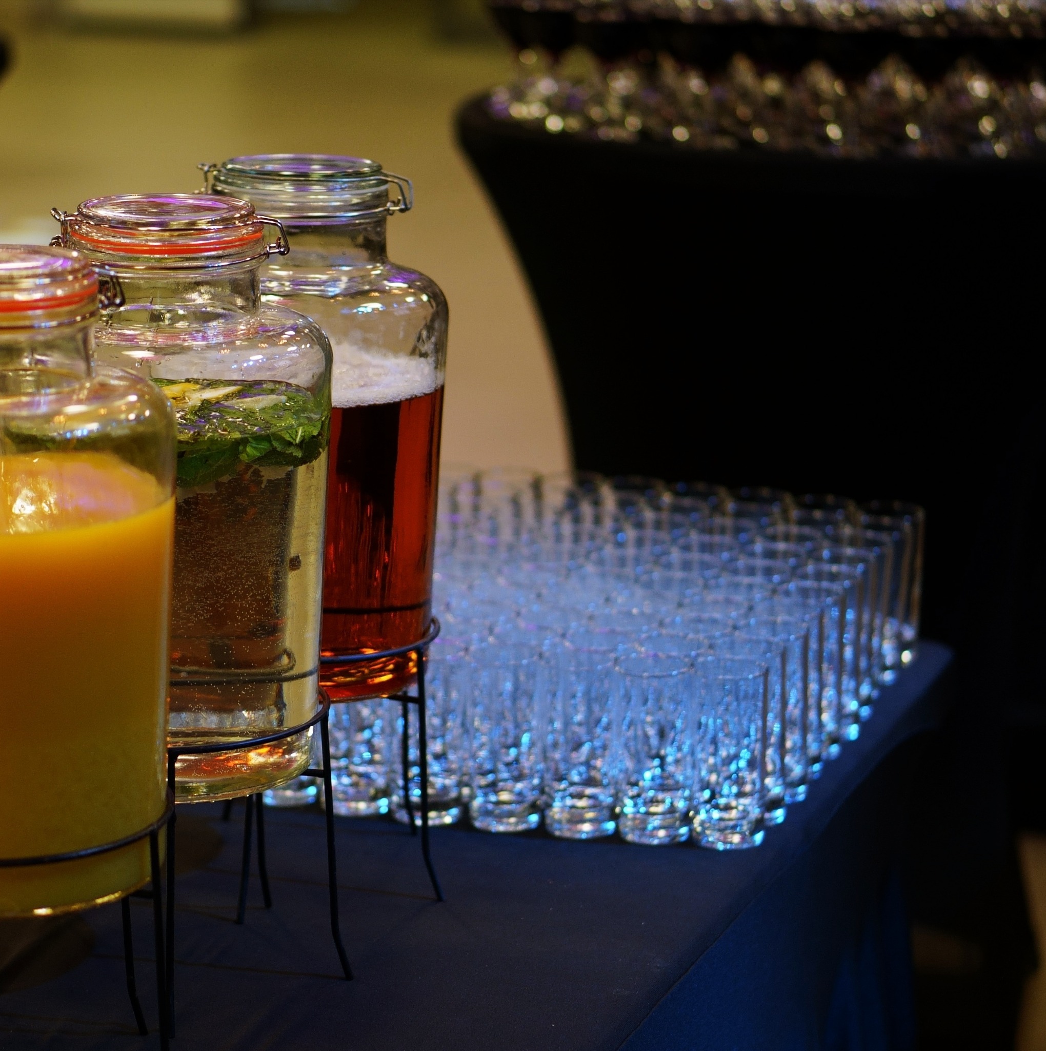 Szklane pojemniki z kranikami na napoje: pomarańczowy sok, woda z miętą i lemonką oraz czerwony napój, ustawione obok rzędów pustych szklanek na ciemnym stole.