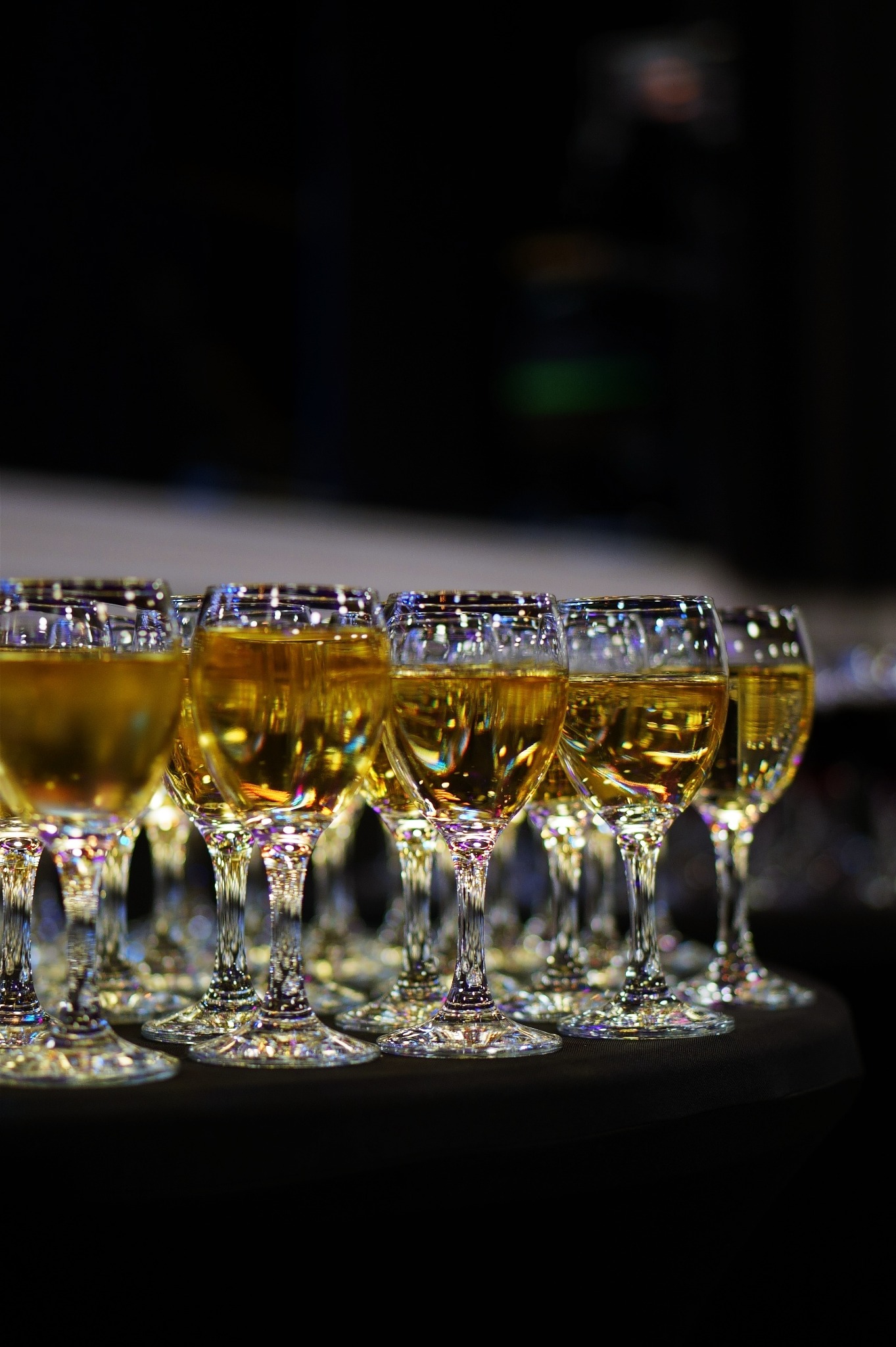 Rząd kieliszków wypełnionych złotym trunkiem, ustawionych na ciemnym tle, z widocznymi refleksami światła na szkle.